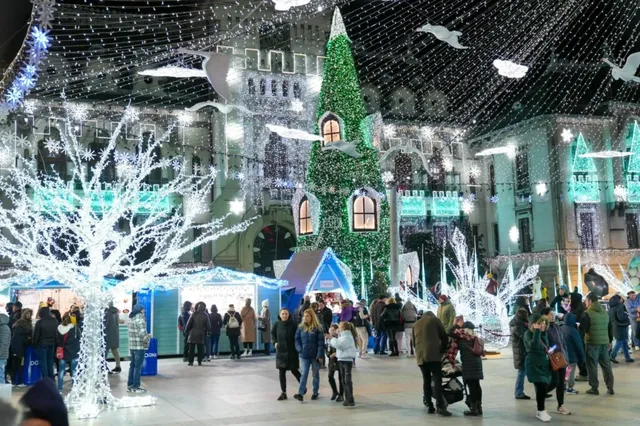 Oamenii stau pe o piață publică noaptea, sub decoruri luminoase de iarnă și un copac de Crăciun înalt decorat cu sclipici verde.