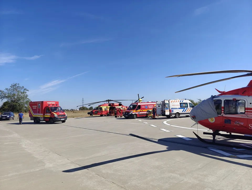 Mai multe ambulanțe și un elicopter roșu sunt parcate pe o suprafață asfaltată sub un cer senin.