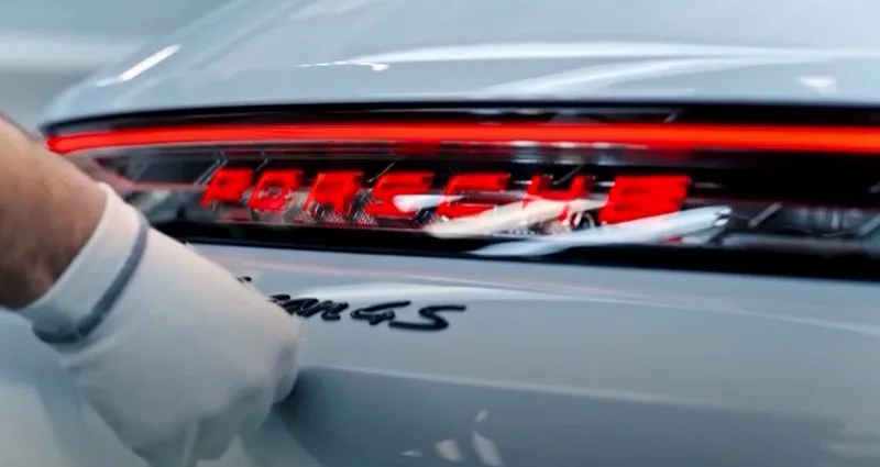 O mână îmbrăcată într-o mănușă albă atinge partea din spate a unei mașini albe, unde este vizibilă inscripția roșie Porsche.