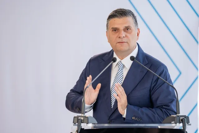 Un bărbat într-un costum albastru și cămașă albă cu cravată albastră stă în spatele unui podium cu două microfoane.