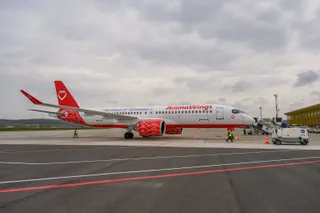 O aeronavă albă cu accente roșii și logo-ul AnimaWings stă pe o pistă de aeroport sub un cer înnorat.