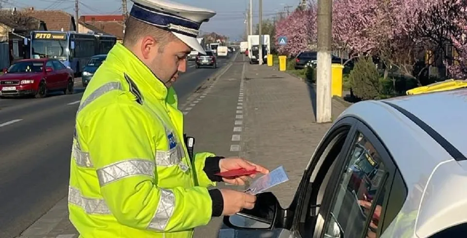 Un agent de poliție în uniformă galbenă reflectorizantă examinează un document oferit de o persoană aflată în interiorul unui autoturism alb.