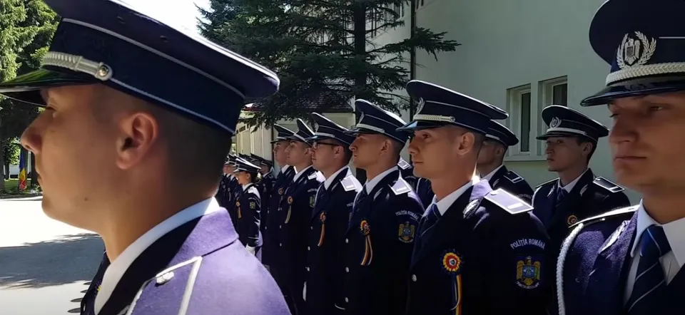O serie de bărbați în uniforme de serviciu și căciuli de poliție stau aliniați în exterior, în fața unei clădiri și a unui copac.
