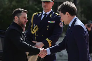 Un bărbat cu barbă și haină neagră strânge mâna unui bărbat într-un costum albastru în fața unui militar în uniformă de gală.