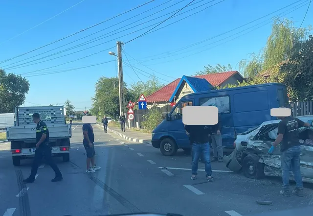 Un autoturism alb avariat stă lângă o autoutilitară albastră pe un drum asfaltat, în prezența câtorva persoane și a unui camion gri.