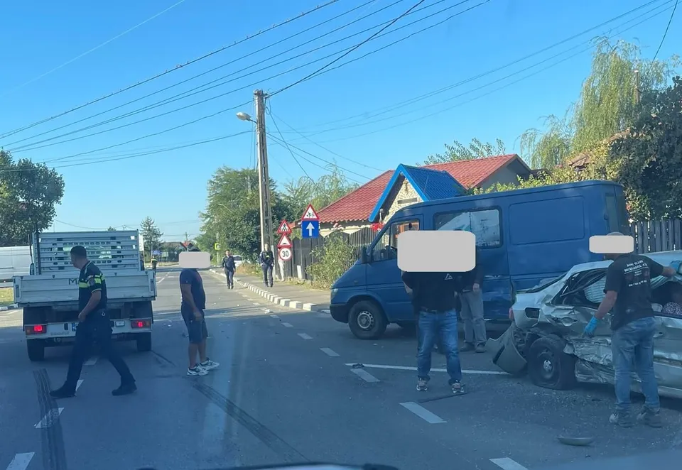 Un autoturism alb avariat stă lângă o autoutilitară albastră pe un drum asfaltat, în prezența câtorva persoane și a unui camion gri.