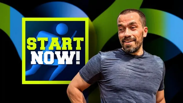 Un bărbat cu barbă și tricou gri stă în fața unui fundal negru cu elemente grafice colorate și un logo dreptunghiular.