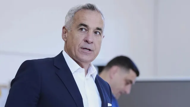 Un bărbat cu părul cărunt, purtând sacou închis la culoare și cămașă albă, privește spre lateral într-un spațiu cu fundal deschis.
