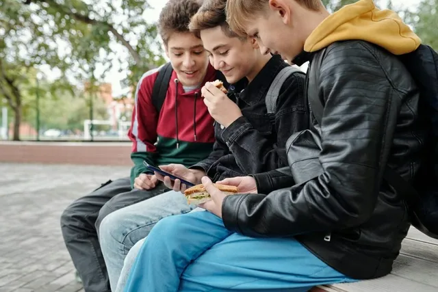 Trei adolescenți stau pe o bancă afară, unul mâncând un sandviș, în timp ce ceilalți privesc un dispozitiv electronic portabil.