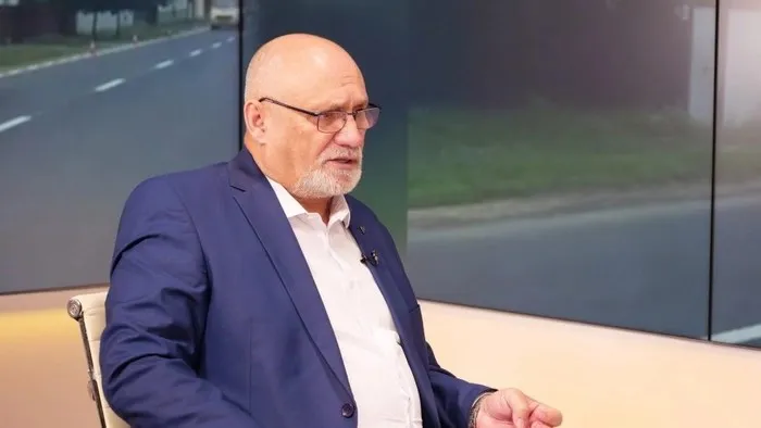 Un bărbat cu ochelari și barbă scurtă poartă sacou albastru și cămașă albă, stând într-un studio cu un fundal de asfalt și iarbă.