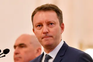 Un bărbat cu părul scurt și costum închis este poziționat în prim-plan, în timp ce un alt bărbat este vizibil în fundal, desfocat.