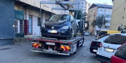 Un autovehicul de culoare închisă este transportat pe platforma unui camion de remorcare pe un drum urban lângă clădiri.