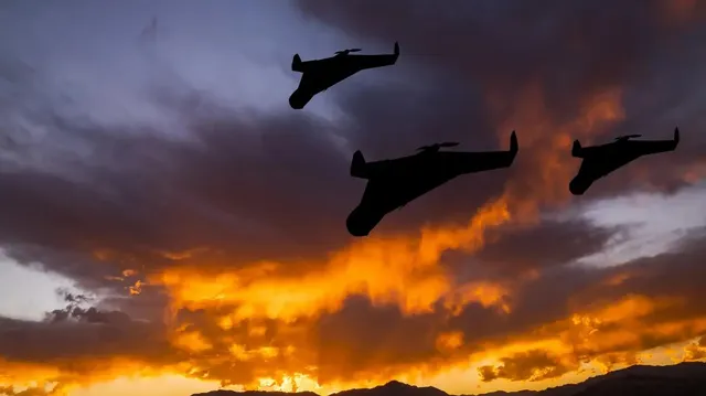 Trei siluete de avioane zboară pe un cer cu nori colorați de lumina apusului.