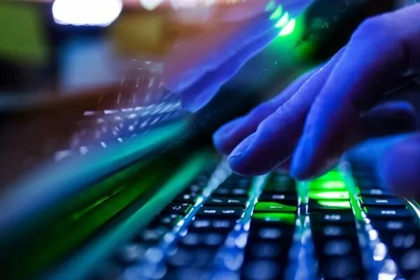 Mâinile unei persoane tastând pe un tastatură iluminată de lumini verzi și albastre într-un mediu întunecat.