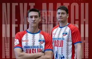 Doi bărbați stau în picioare purtând tricouri albe și roșii cu modele geometrice și inscripții, în fața unui fundal roșu cu textul NEXT MATCH.