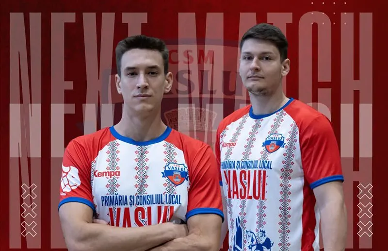 Doi bărbați stau în picioare purtând tricouri albe și roșii cu modele geometrice și inscripții, în fața unui fundal roșu cu textul NEXT MATCH.
