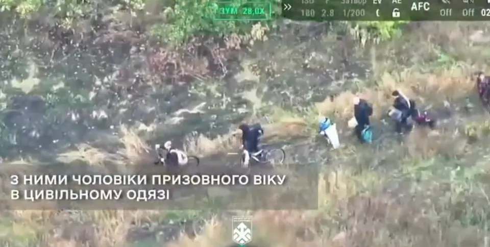 O vedere de sus arată mai multe persoane cu haine colorate deplasându-se printr-o zonă cu vegetație și teren accidentat.