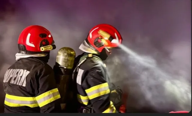Doi pompieri în echipament de protecție și căști roșii folosesc un jet de apă pentru stingerea unui incendiu noaptea.