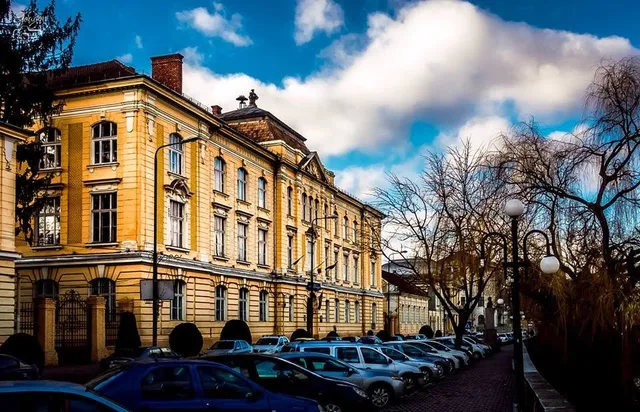 O clădire galbenă cu ferestre multiple este situată lângă un drum unde sunt parcate mai multe mașini sub un cer cu nori.