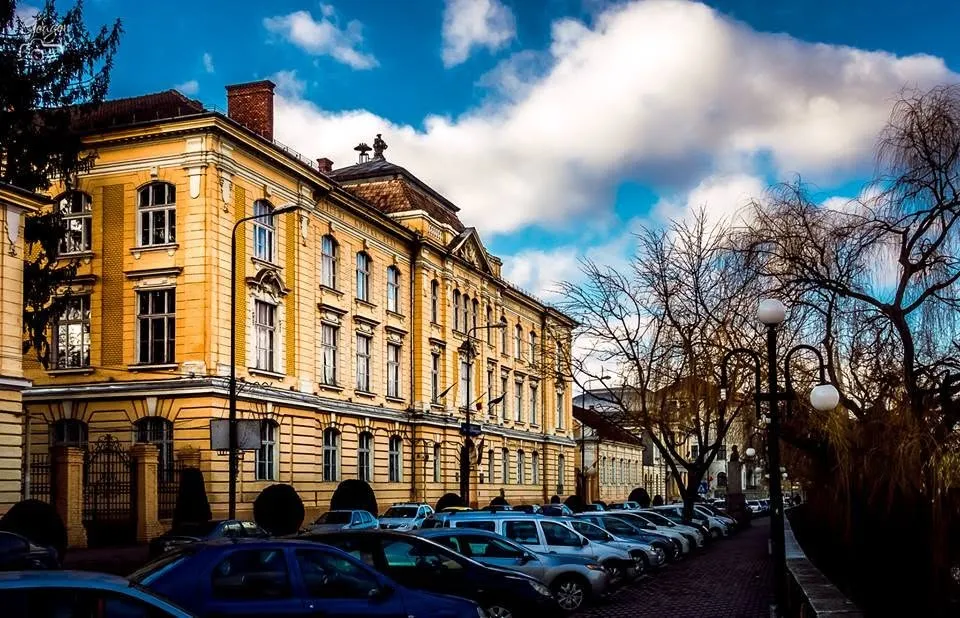 O clădire galbenă cu ferestre multiple este situată lângă un drum unde sunt parcate mai multe mașini sub un cer cu nori.
