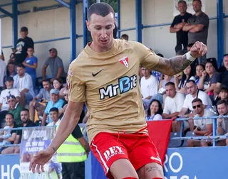 Un bărbat cu tricou auriu și pantaloni scurți roșii aleargă pe un teren de fotbal în fața unor spectatori așezați pe tribune.