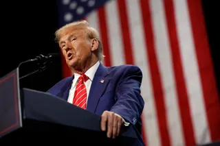 Un bărbat cu părul blond, îmbrăcat în costum albastru și cravată roșie cu dungi albe, vorbește la un podium în fața unui steag american.