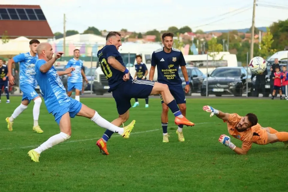 Un jucător de fotbal în echipament albastru lovește mingea pe un teren de iarbă verde în fața unui portar aflat pe sol.