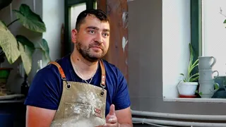 Un bărbat cu barbă și șorț de lucru murdar stă lângă o fereastră lângă plante de interior și câteva vase de ceramică albe.