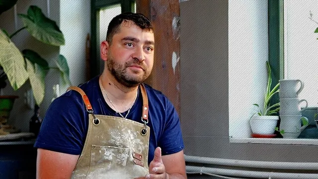 Un bărbat cu barbă și șorț de lucru murdar stă lângă o fereastră lângă plante de interior și câteva vase de ceramică albe.