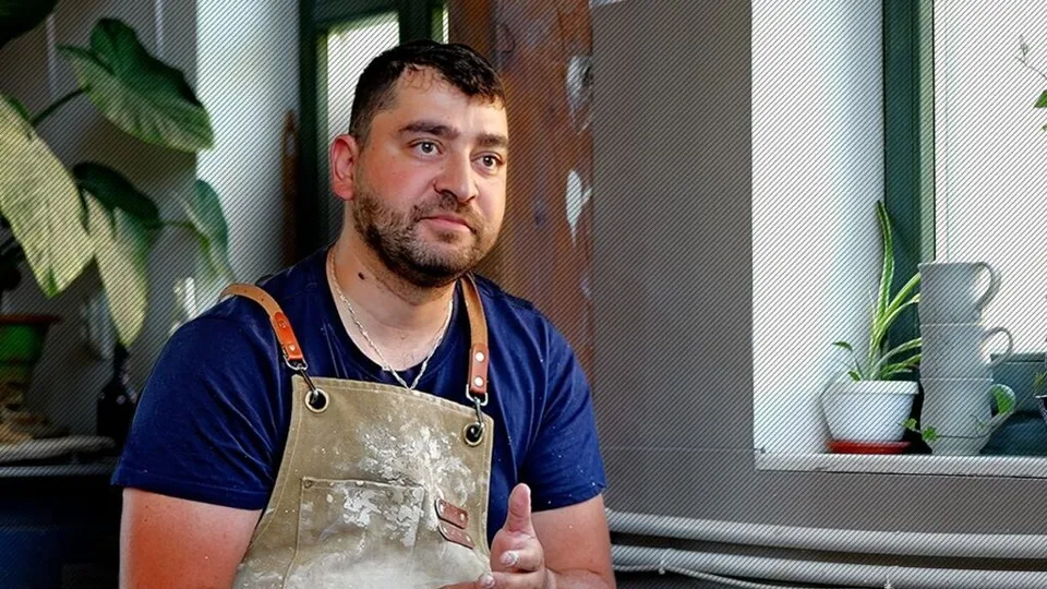 Un bărbat cu barbă și șorț de lucru murdar stă lângă o fereastră lângă plante de interior și câteva vase de ceramică albe.