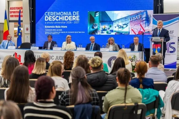 Oamenii stau așezați în rânduri de scaune în fața unei mese lungi pe care se află mai mulți oameni așezați în fața unui ecran mare.