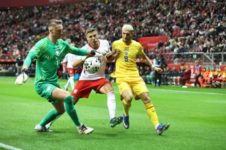 Trei jucători de fotbal concurează pentru un balon pe un teren de iarbă verde în fața unei tribune pline de spectatori.