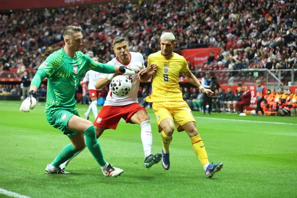Trei jucători de fotbal concurează pentru un balon pe un teren de iarbă verde în fața unei tribune pline de spectatori.