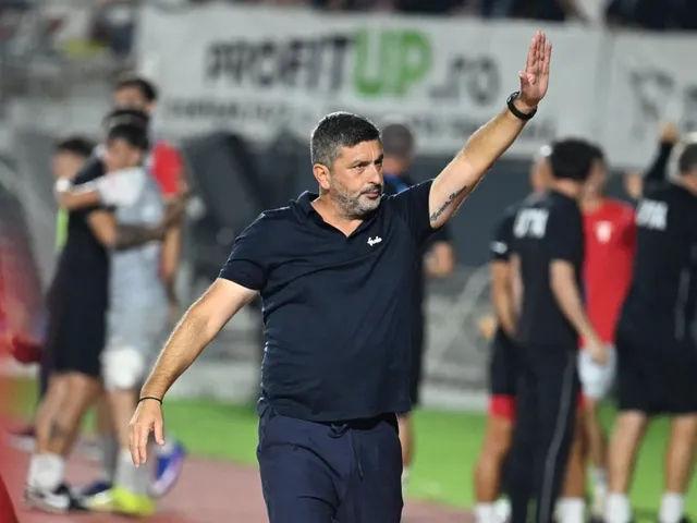 Un bărbat cu barbă, purtând un tricou polo închis la culoare, stă cu un braț ridicat pe un teren de fotbal.