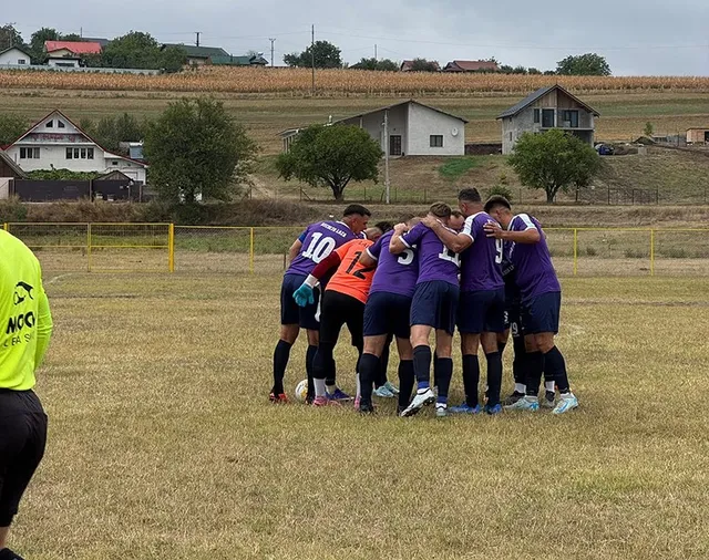Un grup de fotbaliști în echipamente purpurii și portocalii stau îmbrățișați pe un teren de fotbal cu iarbă uscată sub un cer înnorat.