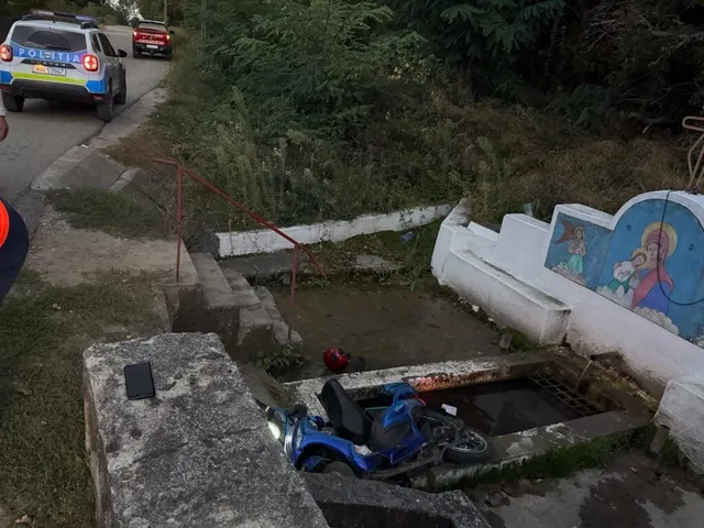 O bicicletă electrică albastră este întinsă pe sol lângă o structură de beton și o treaptă, în timp ce o mașină de poliție este parcată pe drum.