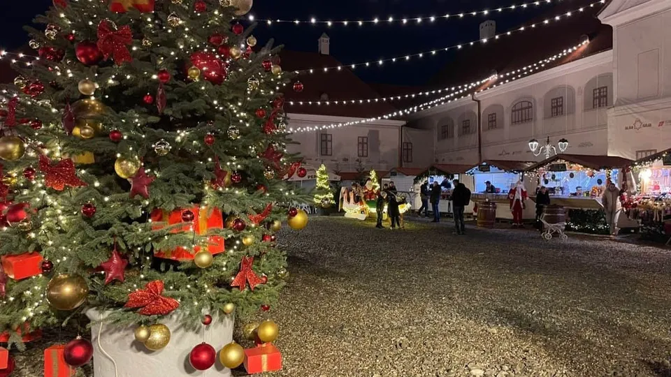 Un brad de Crăciun decorat cu lumini și globuri strălucitoare stă în prim-planul unei piețe noapte, lângă standuri de vânzare și oameni.