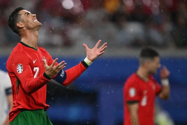 Un bărbat cu tricou roșu și numărul șapte pe piept stă cu capul înspățit și mâinile ridicate pe un teren de fotbal.