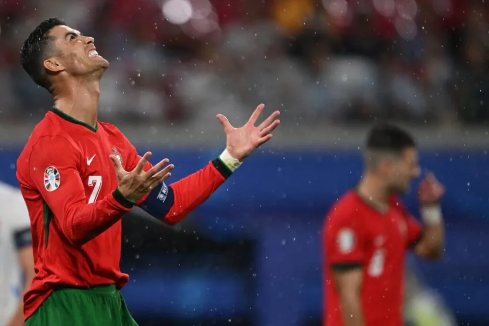 Un bărbat cu tricou roșu și numărul șapte pe piept stă cu capul înspățit și mâinile ridicate pe un teren de fotbal.