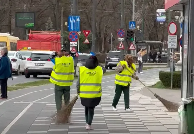 Trei persoane purtând veste fluorescente cu inscripția SALU pe spate folosesc măturători pe un trotuar lângă mașini și semne de circulație stradală.