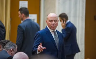 Un bărbat cu pielea deschisă, purtând sacou albastru și cravată cu model, stă în picioare într-o încăpere cu alți bărbați în costume.