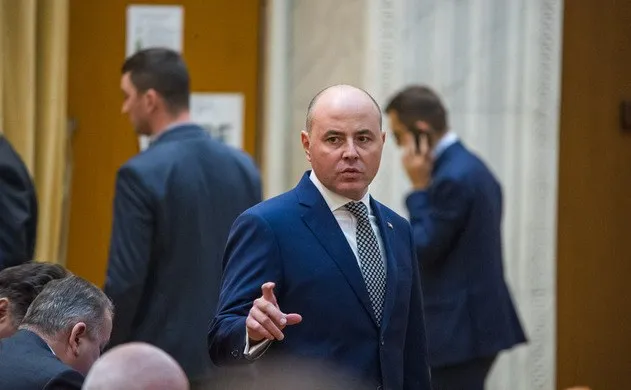 Un bărbat într-un costum albastru și cravată cu model stă în picioare și face un gest cu mâna într-un spațiu interior.