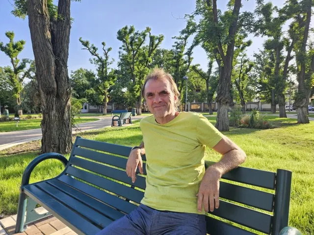 Un bărbat cu tricou galben stă pe o bancă verde într-un parc cu copaci și iarbă verde în fundal.