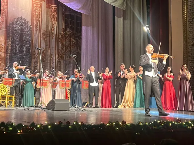 Un grup de muzicieni și artiști în haine de seară stau pe o scenă de teatru în fața unui decor pictat.