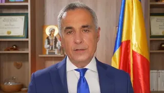 Un bărbat cu părul cărunt, purtând sacou albastru, cămașă albă și cravată albastră, stă în fața unui fundal cu rafturi și un steag.