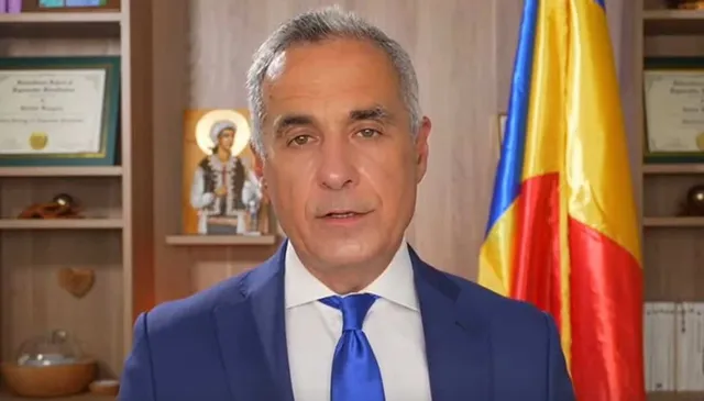 Un bărbat cu părul cărunt, purtând sacou albastru, cămașă albă și cravată albastră, stă în fața unui fundal cu rafturi și un steag.