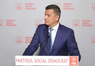 Un bărbat într-un costum albastru și cravată albastră stă în fața unui fundal cu logourile Partidul Social Democrat.