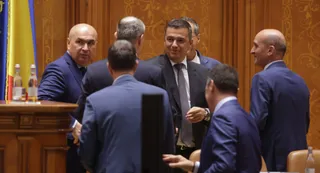 Un grup de bărbați în costume de culoare închisă stau în picioare într-o încăpere cu panouri de lemn și un drapel colorat în fundal.