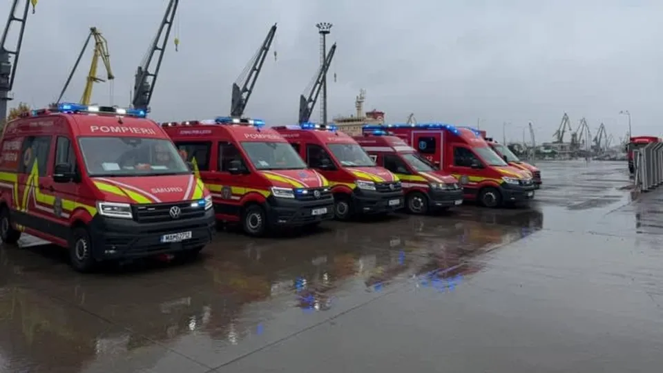 Cinci autovehicule de intervenție roșii cu inscriptiona POMIERI și SMURD, echipate cu faruri și semnalizatoare luminoase, sunt parcate pe un port pavat, cu macazuri industriale în fundal, pe un cer înnorat.