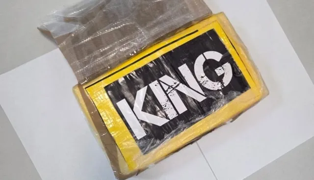 Un pachet dreptunghiular învelit în folie de plastic, cu un etichetă galbenă pe care scrie textul alb "KING" pe un fundal negru, este așezat pe o suprafață albă.